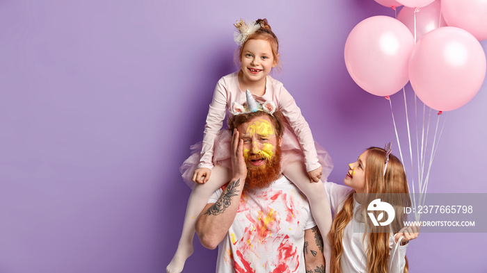 Children and holiday concept. Fatigue bearded dad smeared with paints, gives piggyback to daughter, entrertains children during party, draw and play together, isolated over purple background.