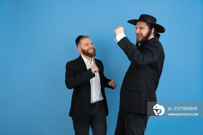 Dancing, having fun. Portrait of a young orthodox jewish men isolated on blue studio background. Purim, business, festival, holiday, celebration Pesach or Passover, judaism, religion concept.