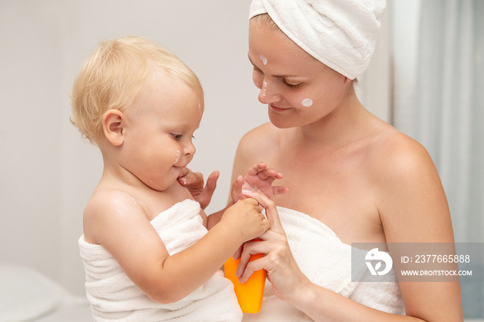 Mother and infant baby in white towels after bathing apply sunscreen or after sun lotion or cream. Children skin care in a hotel or bedroom. Spf, skin moisturizer, vacation concept