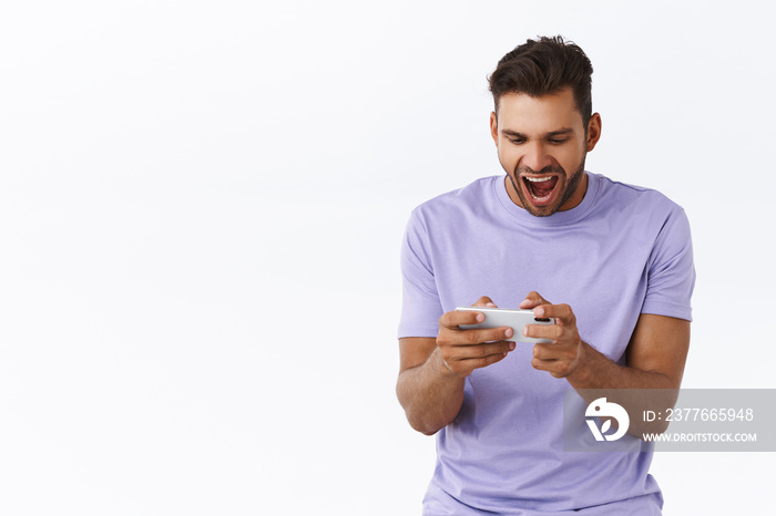 Cheerful, excited good-looking hispanic bearded guy in purple t-shirt, looking astonished and thrilled, playing awesome new smartphone game, yelling and smiling pleased, winning, white background