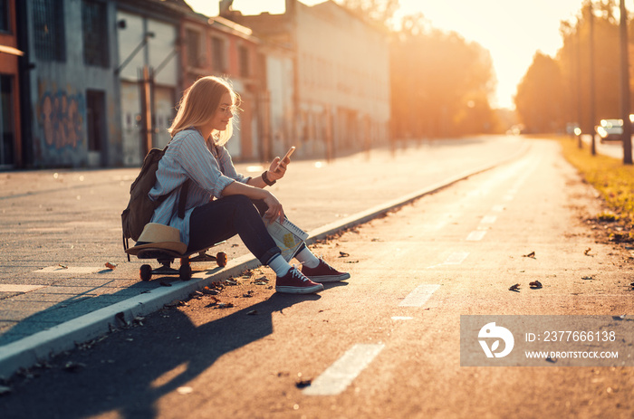 Smiling girl sitting on long board in the city during sunset and looking at phone.