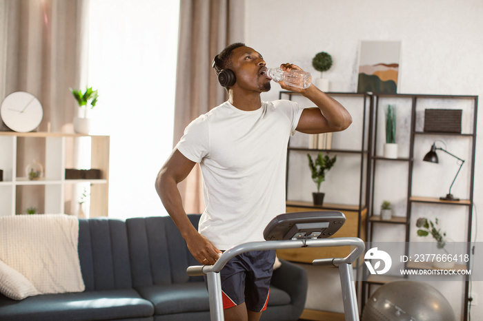 Mature sportsman in white t-shirt running on treadmill at home listening to music in headphones and drinking water from bottle. African American athletic man stays hydrated while working out