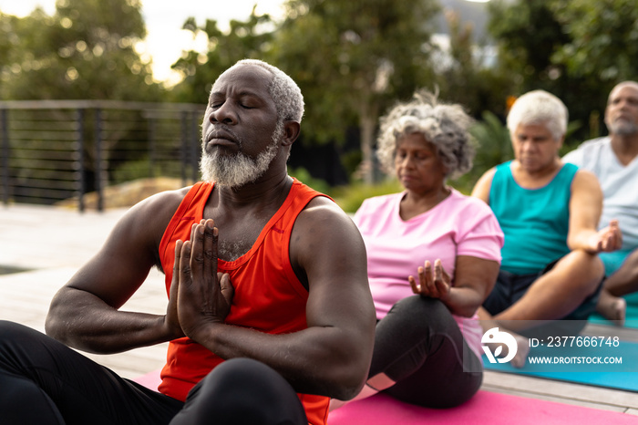 Senior multiracial friends meditating while sitting on mats against plants in yard at nursing home