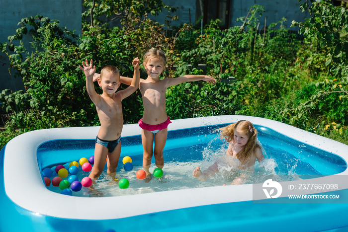 Three happy friends are splashing in an inflatable pool in the garden. Concept of summer season and recreation. Kids having fun together. Children playing and swimming in water on the backyard.