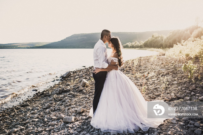 groom and bride in wedding dress stand in embrace on rocky bank near river in summer outdoors In rays of setting sun. Bakota, Ukraine