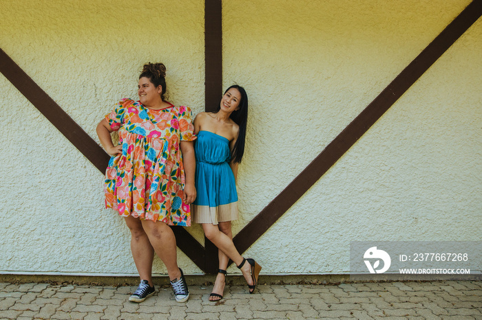 2 friends wearing dresses stand against a wall