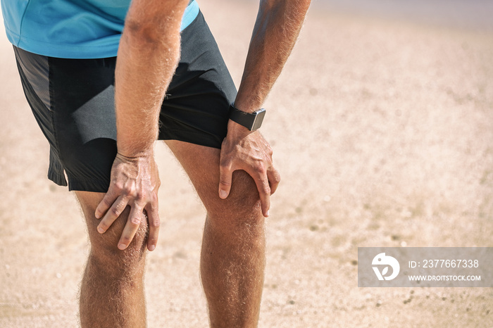 Male runner resting taking a break tired with knee pain after beach jog wearing smartwatch wearable technology for exercise monitoring weight loss.