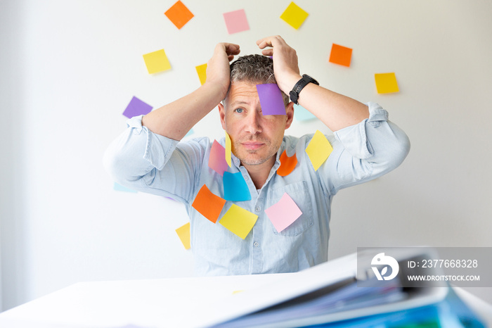 Man sits behind white office desk in front of wall with many colorful notes and also has on himself stick many notes and is looking for an idea