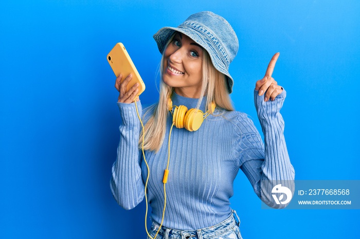 Young blonde woman using smartphone wearing headphones surprised with an idea or question pointing finger with happy face, number one