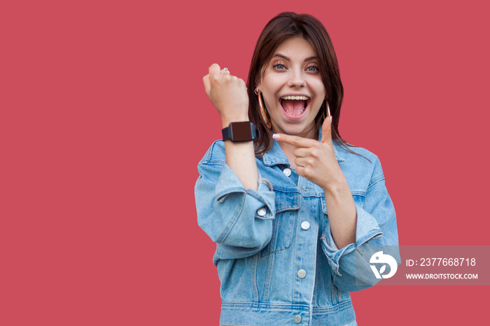 Portrait of surprised happy beautiful brunette young woman in denim casual style standing showing and pointing at screen of her smart watch with shocked face. studio shot, isolated on red background.