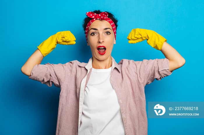 Strong young woman showing biceps in cleaning gloves on blue background.