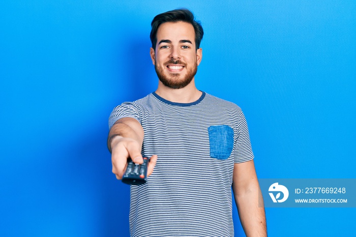 Handsome caucasian man with beard holding television remote control smiling with a happy and cool smile on face. showing teeth.