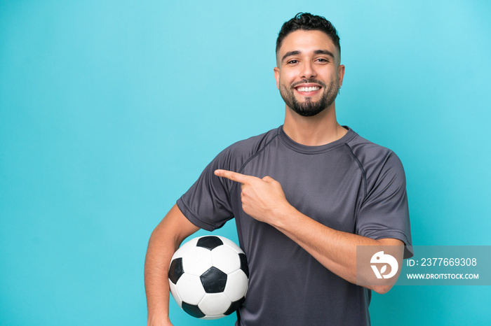 Young Arab handsome man isolated on blue background with soccer ball and pointing to the lateral