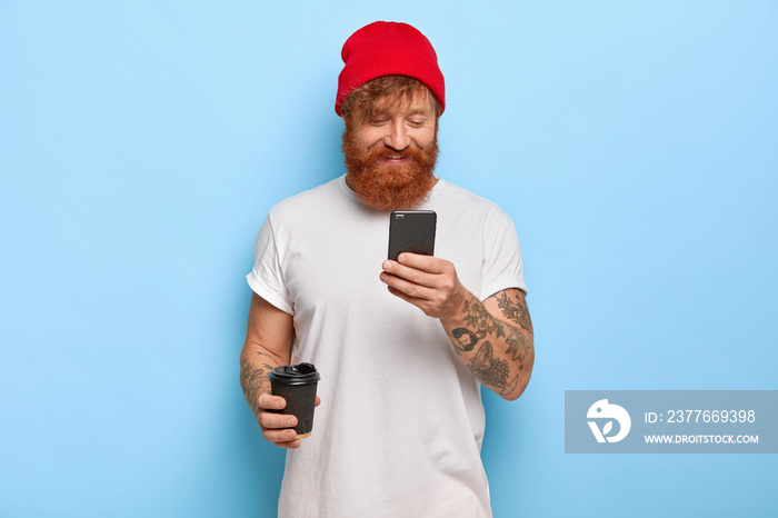 Positive emotions and modern technologies concept. Cheerful stylish man wears red hat and white t shirt, has ginger beard chats with friends via cell phone connected to wireless internet drinks coffee