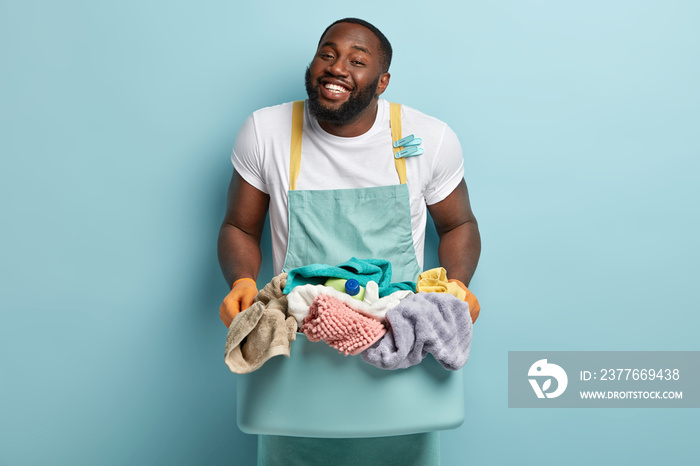 Friendly looking glad smiling dark skinned man with stubble, white teeth, holds basin with linen for washing, being in high spirit, wears casual white t shirt and apron, models over blue background