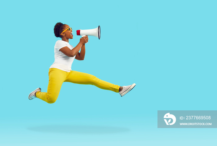 Happy young woman shouts through megaphone. Funny crazy Afro American lady in T shirt, yellow pants and sunglasses jumps high in air and yells in loudspeaker isolated on bright blue color background