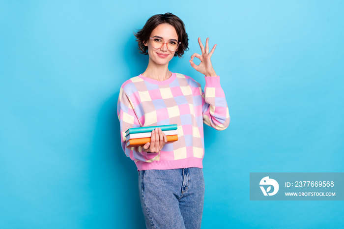 Photo of cute millennial brunette lady hold book show okey wear spectacles checkered pullover jeans isolated on blue background