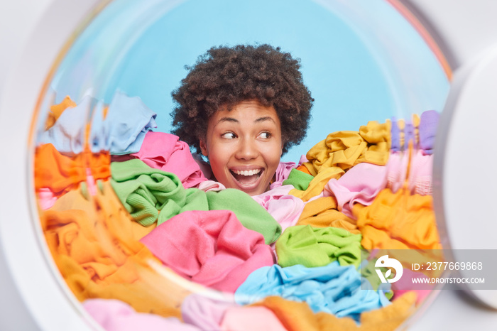 Head of funny woman with cheerful expression stuck in heap of mutlicolored laundry blue background. View from inside of washer. Busy housewife has curly hair does laundering at home. Housework