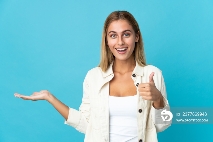 Young blonde woman isolated on blue background holding copyspace imaginary on the palm to insert an ad and with thumbs up