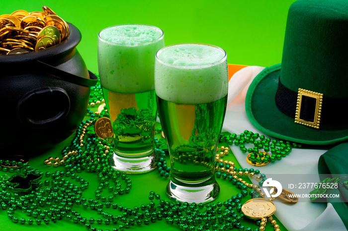 The luck of the Irish meme and Happy St Patricks day concept theme with two glasses of dyed beer, leprechaun hat, beads necklace and pot of gold coins on the Ireland flag isolated on green background