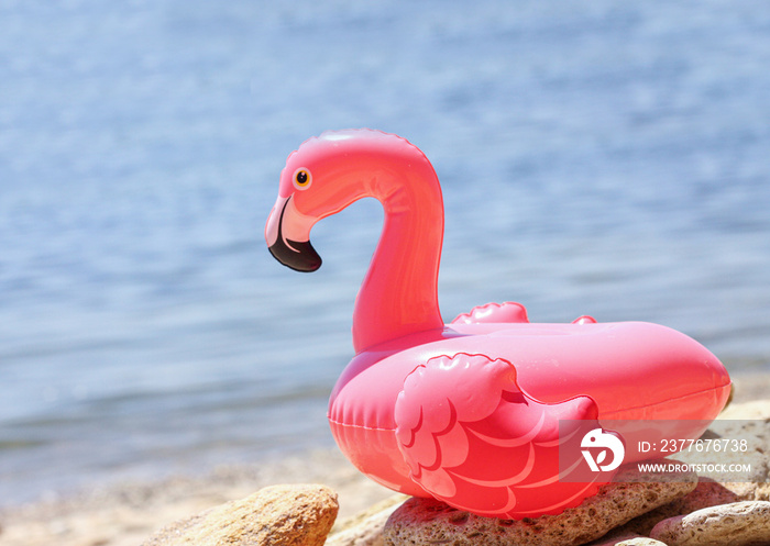 Inflatable little pink flamingo by the sea on  pebbles. Recreation and vacation concept, children’s game and fun. Summer relaxation on the beach. The sun is shining brightly, weather is great