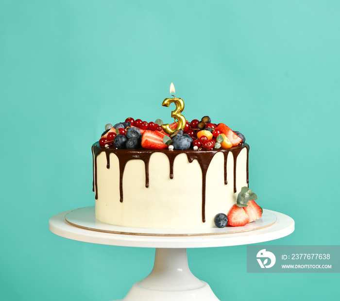 Three years old Birthday party sweet cake with chocolate drips and fresh fruits Raspberry strawberry blueberry cranberry. 3 lighted candle on popular blue mint background