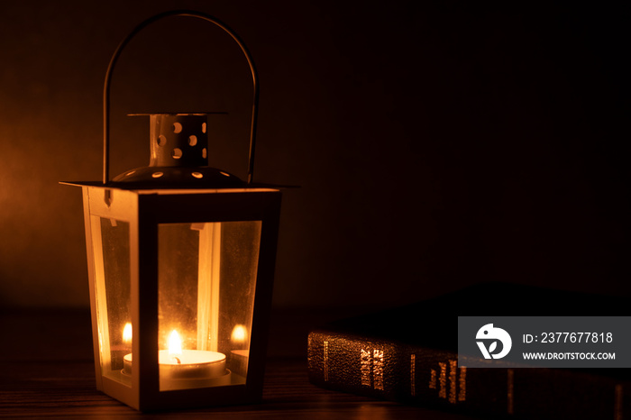 An old lamp illuminated by a small candle flame with a closed Holy Bible Book with golden text on a dark background with copy space. Word of God is a light to the world biblical concept. A closeup.