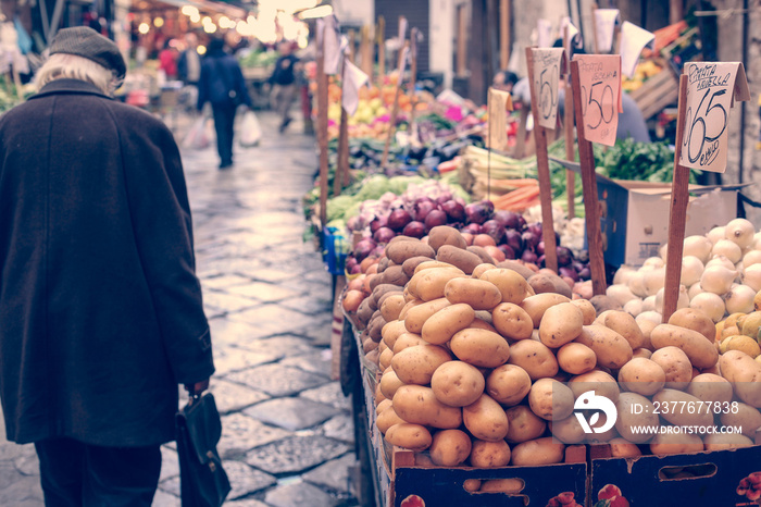 Food market in Palermo, sicily, with a pile of raw potatoes in the foreground, with an old man walking past the market. Prices visible next to food.