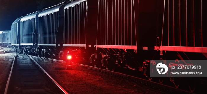 Large cargo train with goods leaving the terminal at night, wagons (containers) close-up. Concept urban scene. Illumination, red light. Freight transportation, industry, business, logistics, delivery