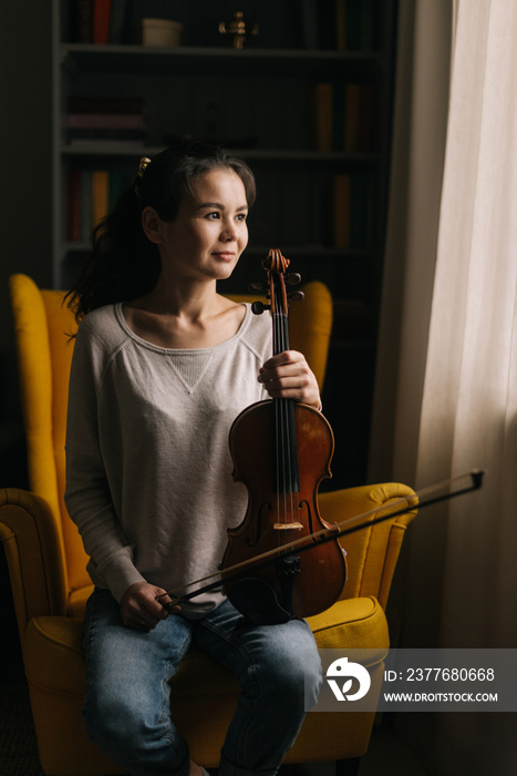 Beautiful young woman posing with a violin in her hand, sitting on a chair in a room with a modern interior. Girl musician is posing with musical instrument