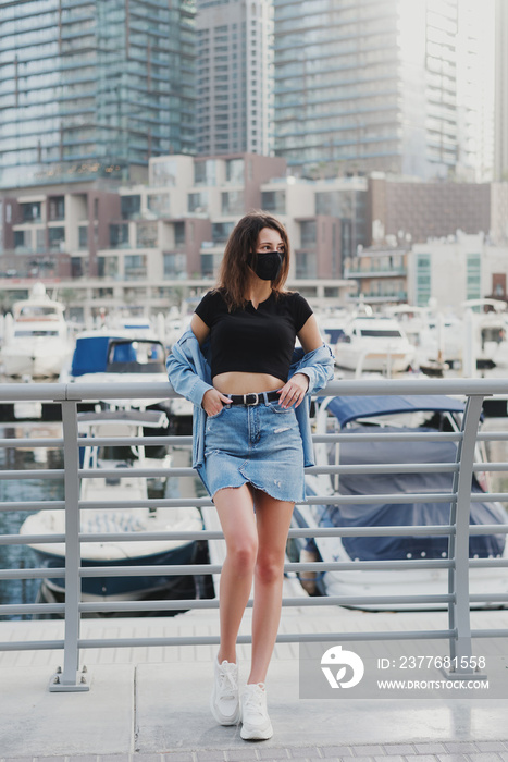 Young woman in short denim skirt wearing reusable face mask of black color and standing at city street. New normal during Covid 19 pandemic