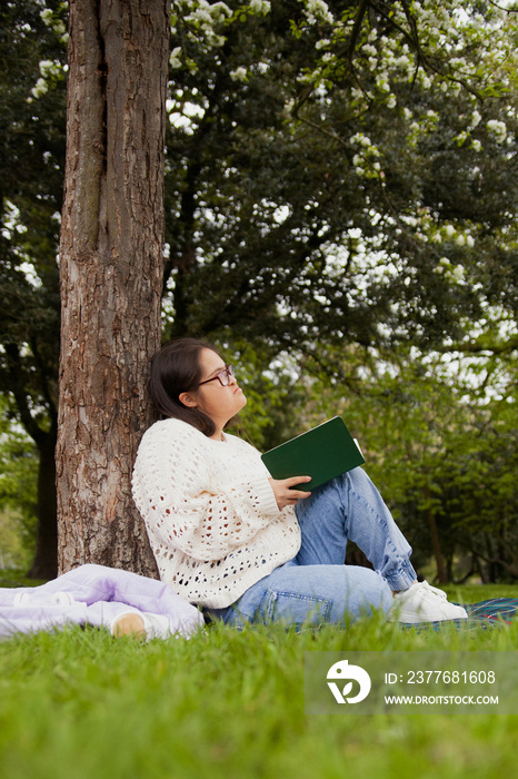 Curvy woman with Down syndrome reading and daydreaming in the park