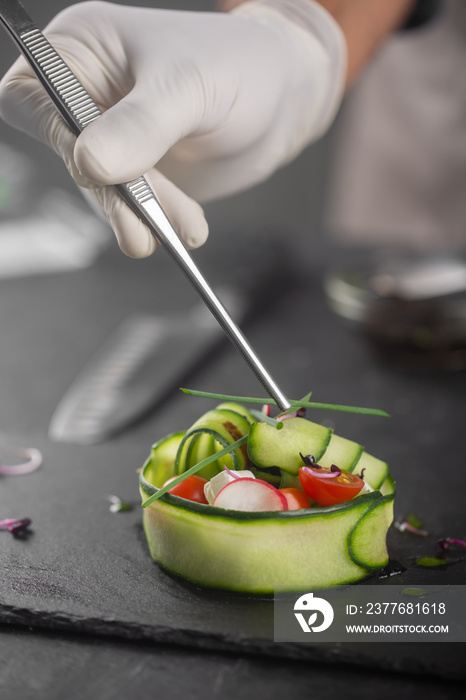 A vegetable snack made from fresh vegetables such as cucumber, tomato, feta cheese and microgreens. A gloved chef spreads green onions with tweezers on a gourmet platter