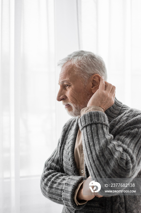 senior and thoughtful man in knitted cardigan looking away near window while suffering from alzheimer syndrome.