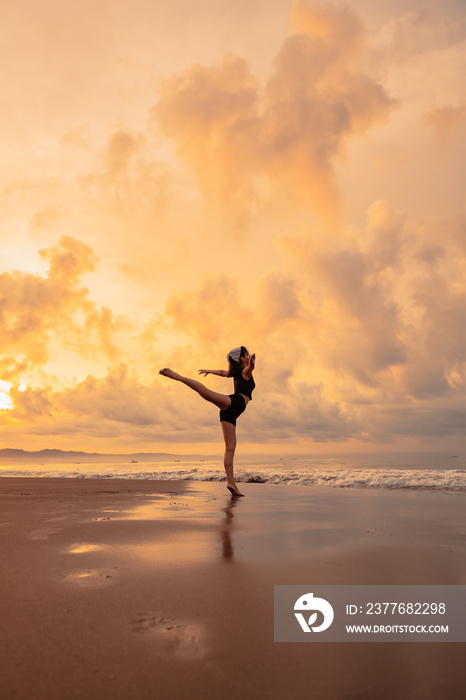 A ballerina in a black dress is practicing ballet moves on the beach with very flexible movements with a view of the clouds behind