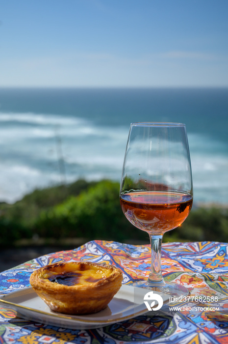 Portugal’s food and drink, glass of porto wine or muscatel de setubal and sweet dessert Pastel de nata egg custard tart pastry served with view on blue Atlantic ocean in Lisbon area, Portugal