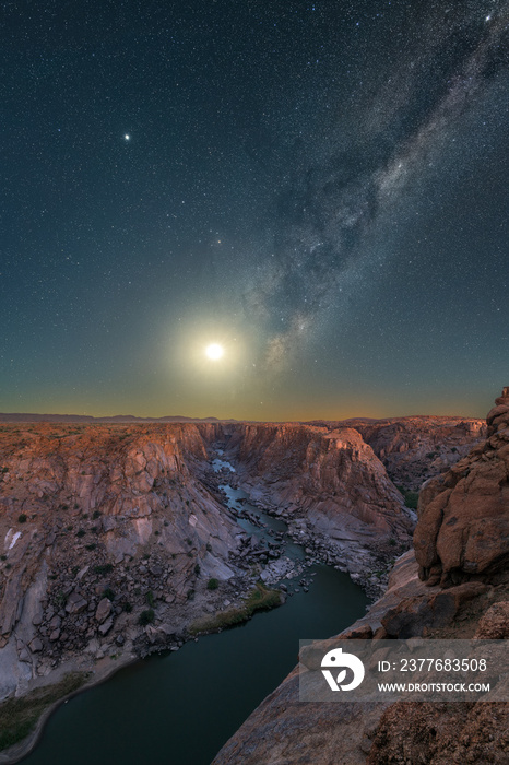 A majestic vertical night sky photograph with the Moon rising below the Milky Way in the deep blue sky, with views of the river and mountains at the Augrabies Falls National Park in South Africa.