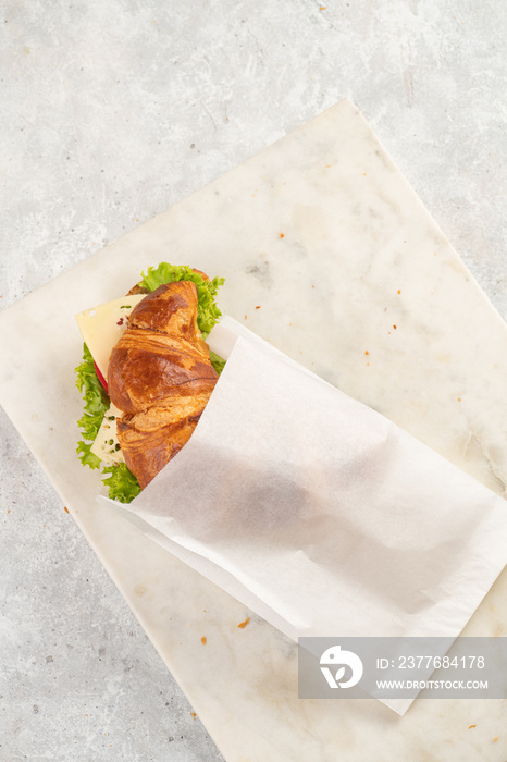 lye croissant sandwich with iberian ham, tomato slices, lettuce and cheese in a breakfast white paper bag on marble board on grey background