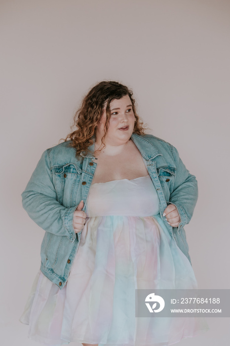 portrait of a plus size woman wearing a denim jacket against a pink background