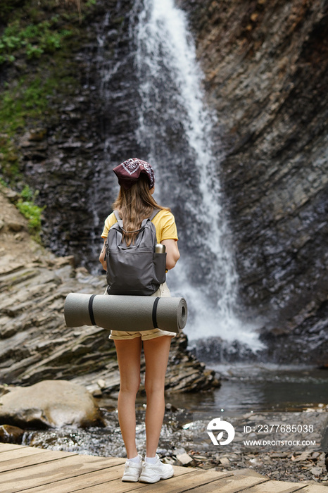 Vertical image of stylishly dressed young female tourist with backpack and mat looking at waterfall and rocks while on vacation. The concept of communing with nature and traveling with camping