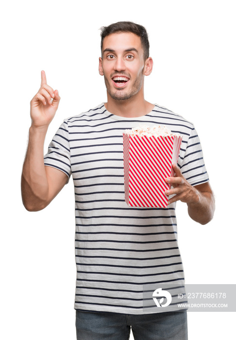 Handsome young man eating popcorn surprised with an idea or question pointing finger with happy face, number one