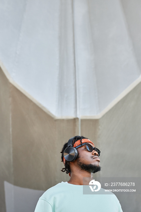 Minimal portrait of young black man wearing headphones standing by concrete wall outdoors and looking up, copy space