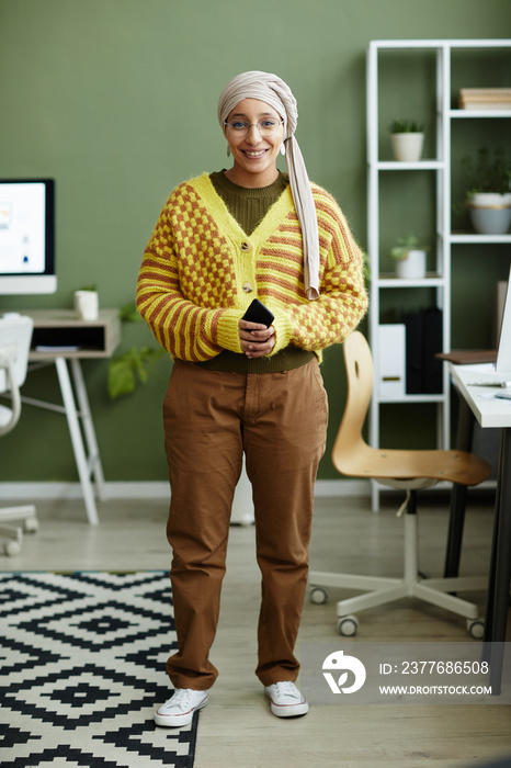 Full length portrait of Middle Eastern young woman wearing vibrant pattern and smiling at camera while standing in office