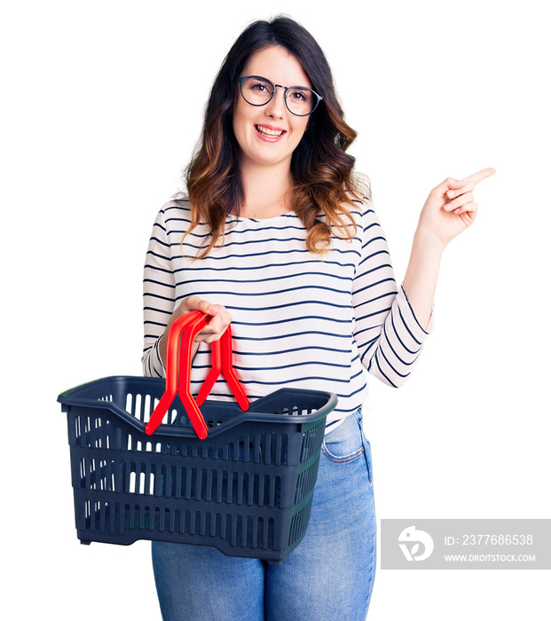 Beautiful young brunette woman holding supermarket shopping basket smiling happy pointing with hand and finger to the side