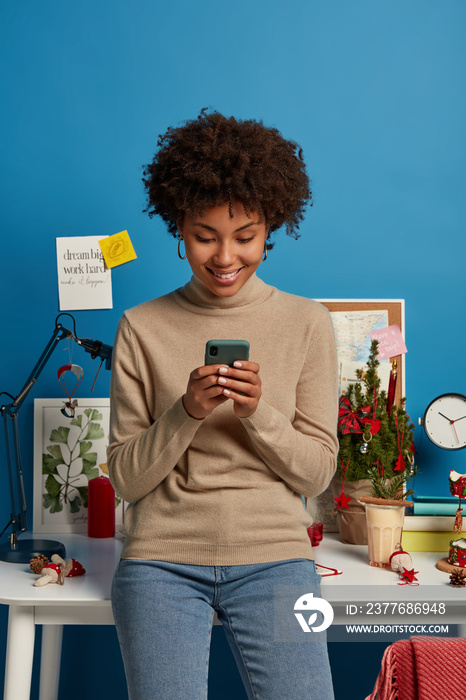 Positive ethnic woman blogger reads comments in blog, enjoys online communication, uses modern cellular, stands in coworking space, wears turtleneck and jeans, watches video in social networks