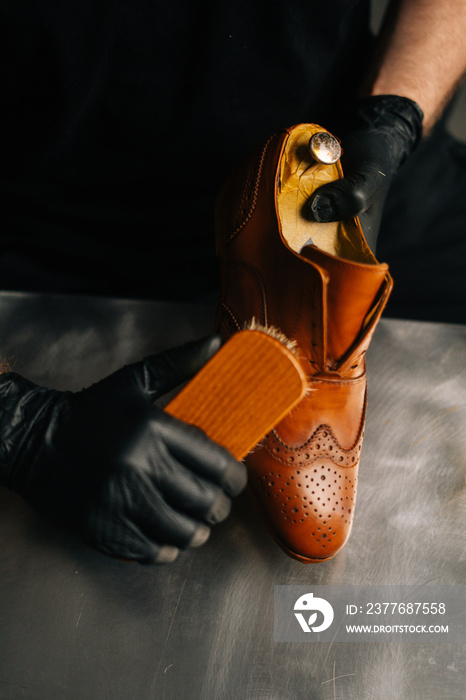 Close-up view of shoemaker in black gloves polishing light brown leather shoes with brush during restoration working. Concept of cobbler artisan repairing and restoration work in shoe repair shop.