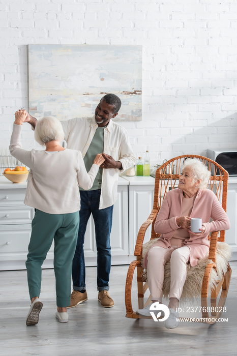 Positive multiethnic pensioners dancing near senior friend in rocking chair in nursing home