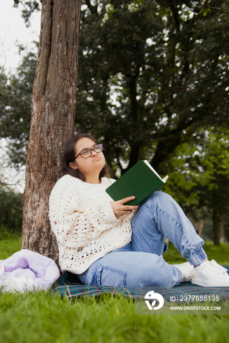 Curvy woman with Down syndrome reading and daydreaming in the park