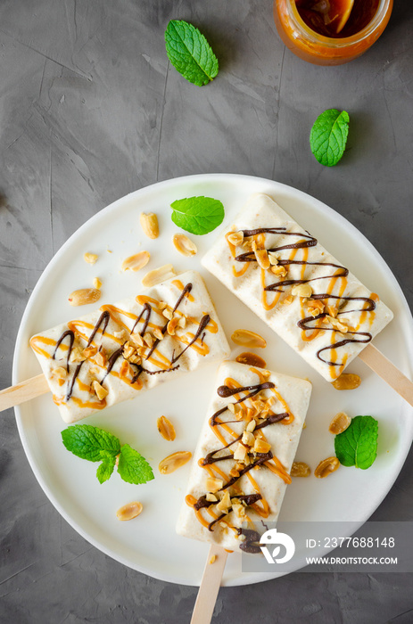 Homemade ice cream with nougat. caramel and peanuts on a stick with mint leaves on a white plate on a gray concrete background. Vertical, top view.