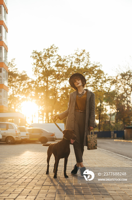 Stylish lady in a coat and hat walking a dog on a leash in the autumn evening at sunset. Portrait of a fashionable girl walking her pet pet down the street. Vertical